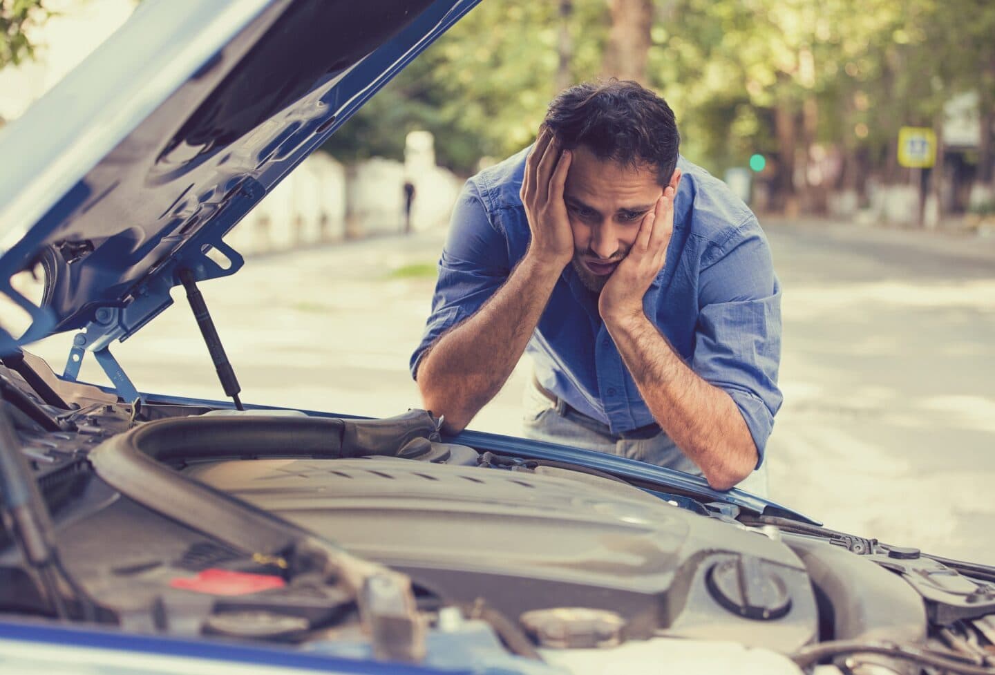 A man appears stressed while looking at his car's open hood on the side of a tree-lined street.